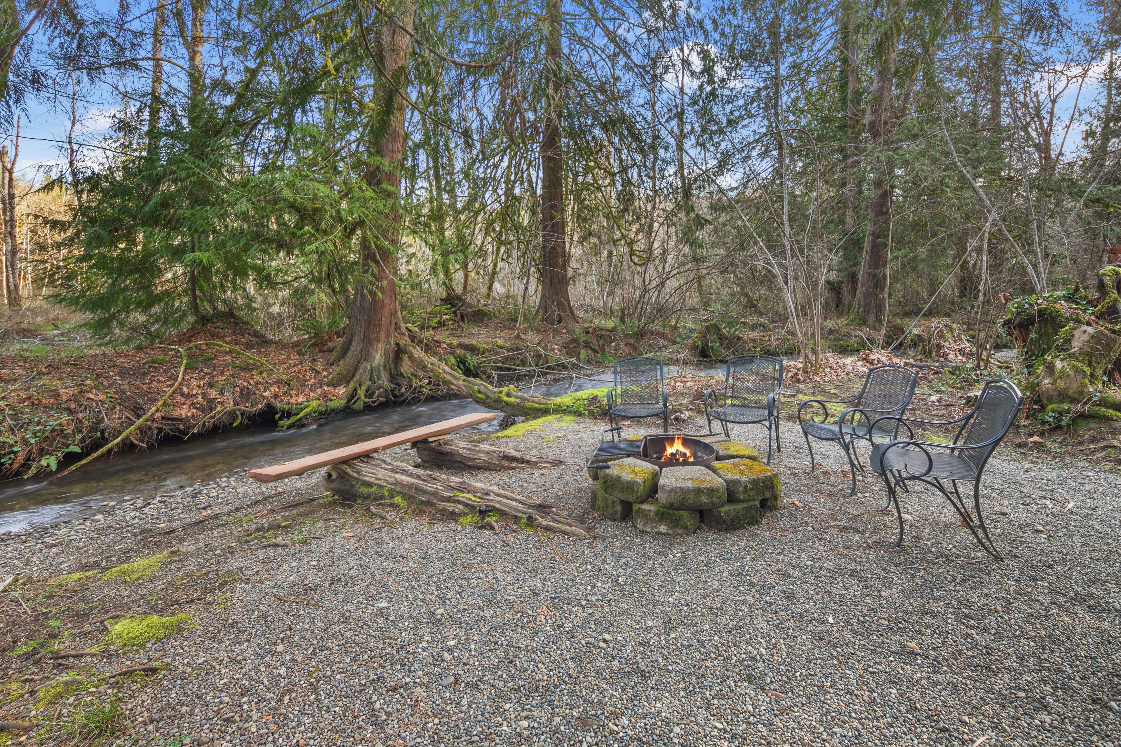 A forested campsite with a gravel area, a fire pit, and metal chairs.