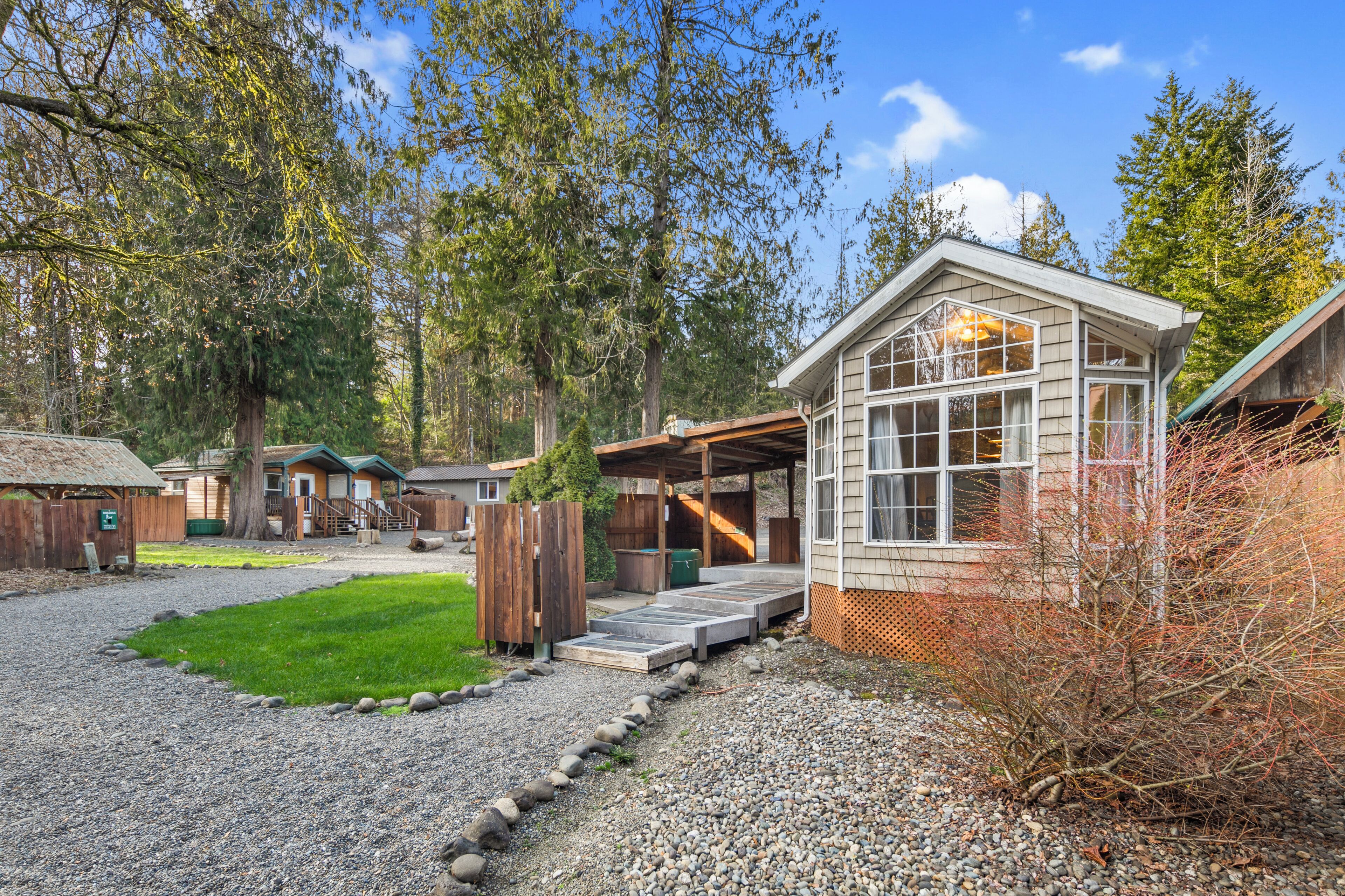 A rustic cabin-style building with a covered porch and a small deck, surrounded by a gravel driveway and a grassy area.