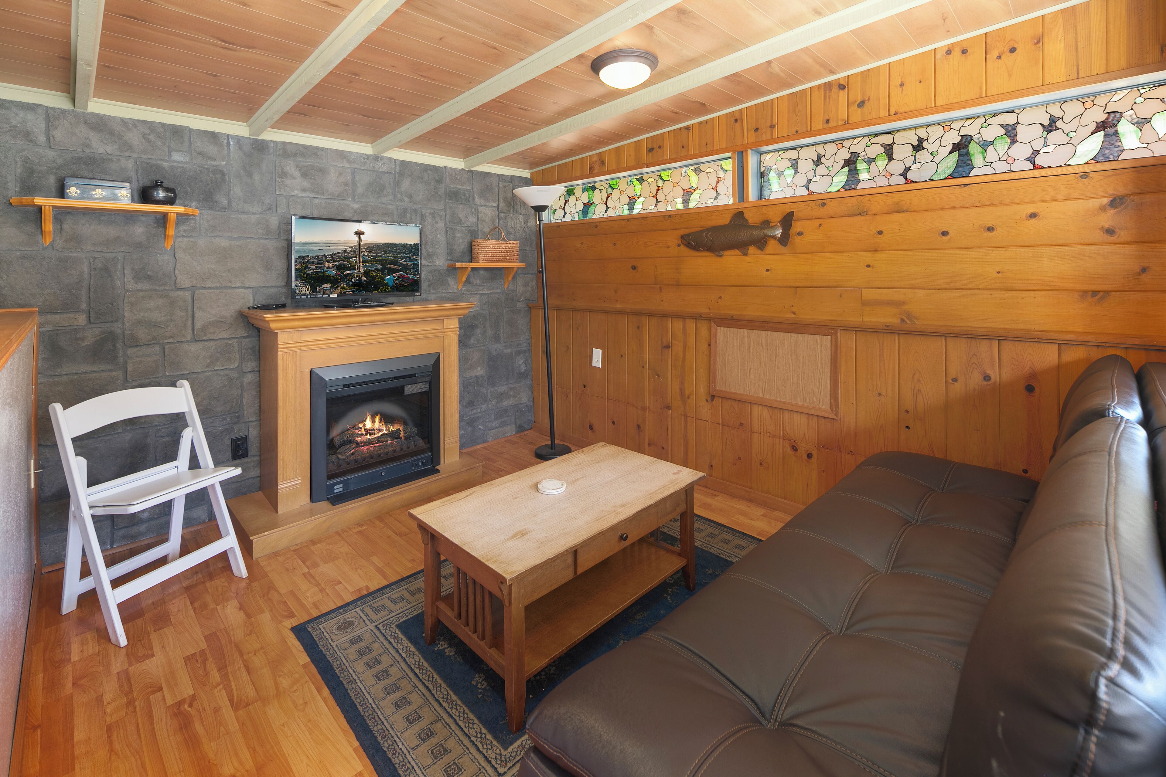 A cozy living room with a fireplace, wooden walls, a stone accent wall, and a large window with a view.