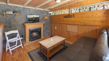 A cozy living room with a fireplace, wooden walls, a stone accent wall, and a large window with a view.