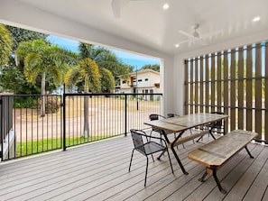 A covered outdoor area with a wooden deck, a table with chairs, and a view of a residential street with palm trees. - Picnic Point (Picnic Bay)