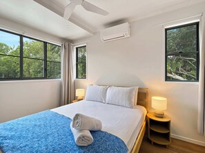 Habitación de hotel con cama, dos mesitas de noche con lámparas, ventilador de techo, equipo de aire acondicionado y ventanas amplias con cortinas.