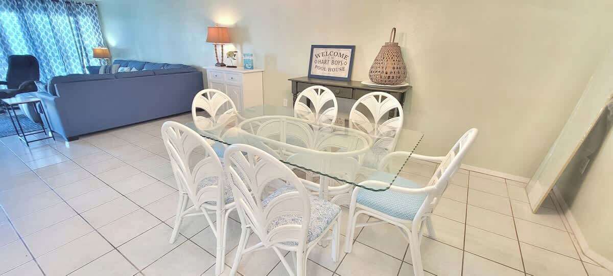 A dining area with a glass table and white chairs, a sofa, a lamp, and a sign that reads "Welcome Hart Boys Pool House".
