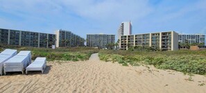 A beachfront hotel with multiple high-rise buildings, a sandy path leading to the beach, and stacks of white furniture.