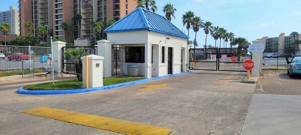 Une entrée fermée par un portail, avec un bâtiment surmonté d’un toit bleu, un panneau de signalisation et une zone de stationnement délimitée par des lignes jaunes.