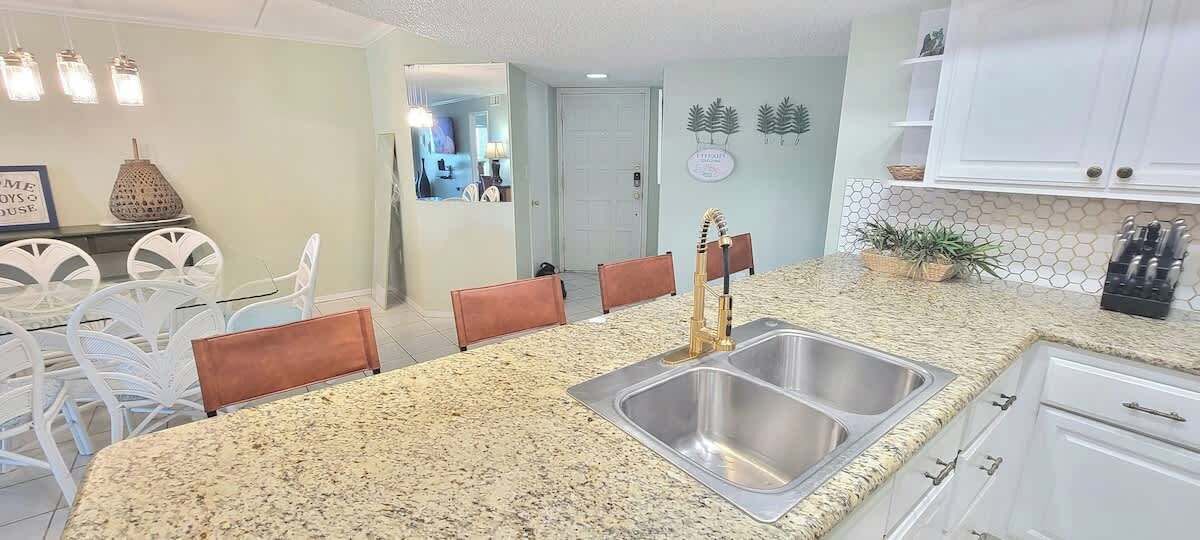 Cocina con encimeras de granito, pileta de acero inoxidable, gabinetes blancos y una zona de comedor con sillas blancas y un banco marrón.