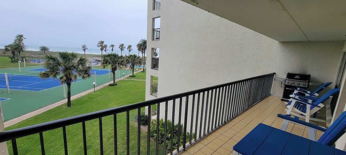 A balcony with a view of a tennis court and palm trees, a grill, and blue chairs.