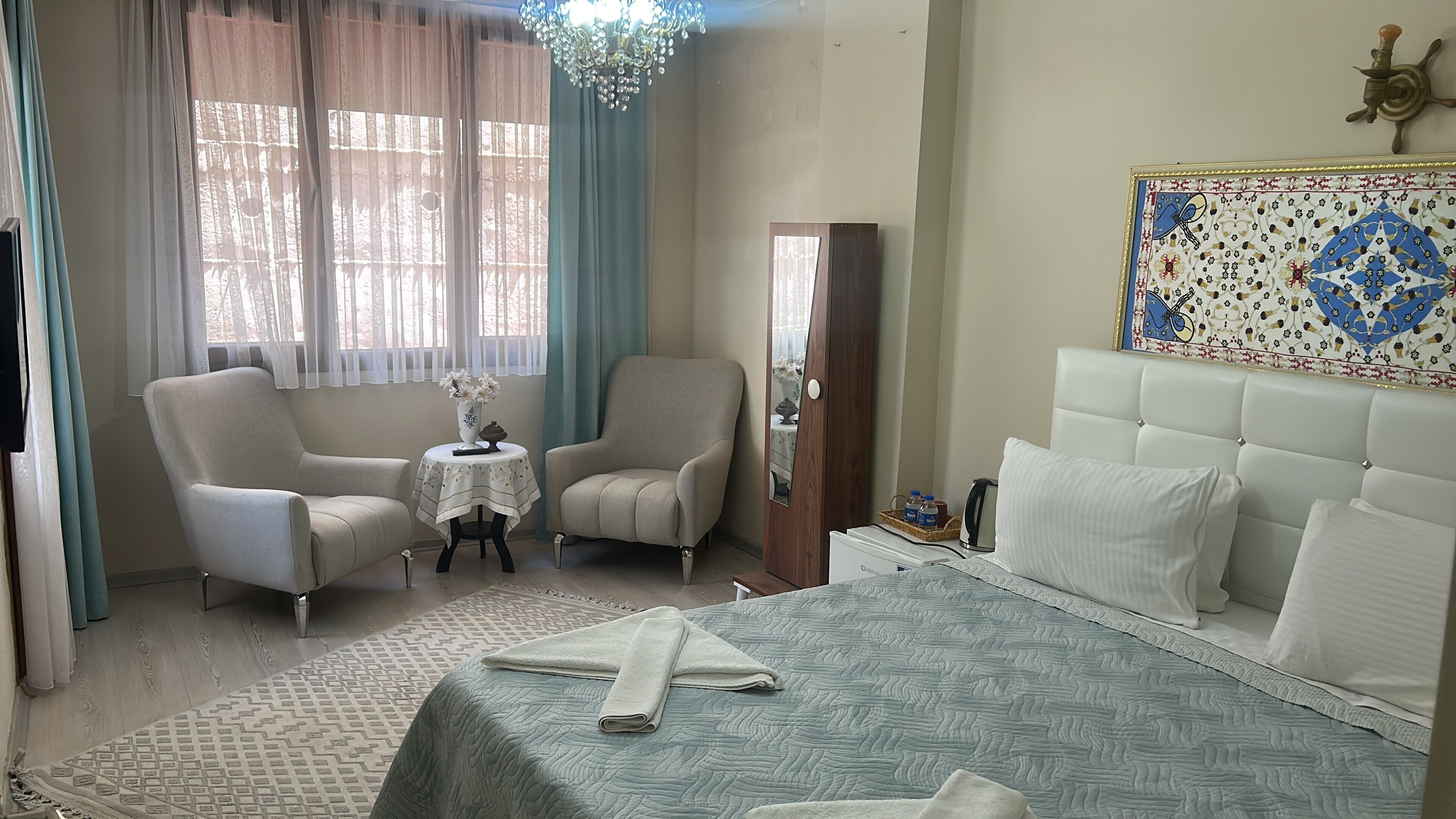 Ein Hotelzimmer mit einem Bett, zwei Sesseln, einem kleinen Tisch mit Blumenvase, einem Spiegel und einer Wanddekoration.