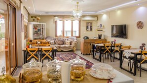 A spacious living room with a large sofa, dining area with wooden chairs, a chandelier, and a TV mounted on the wall. - Ephesus Palace (Selçuk)