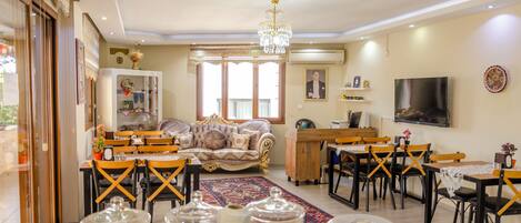 A spacious living room with a large sofa, dining area with wooden chairs, a chandelier, and a TV mounted on the wall.