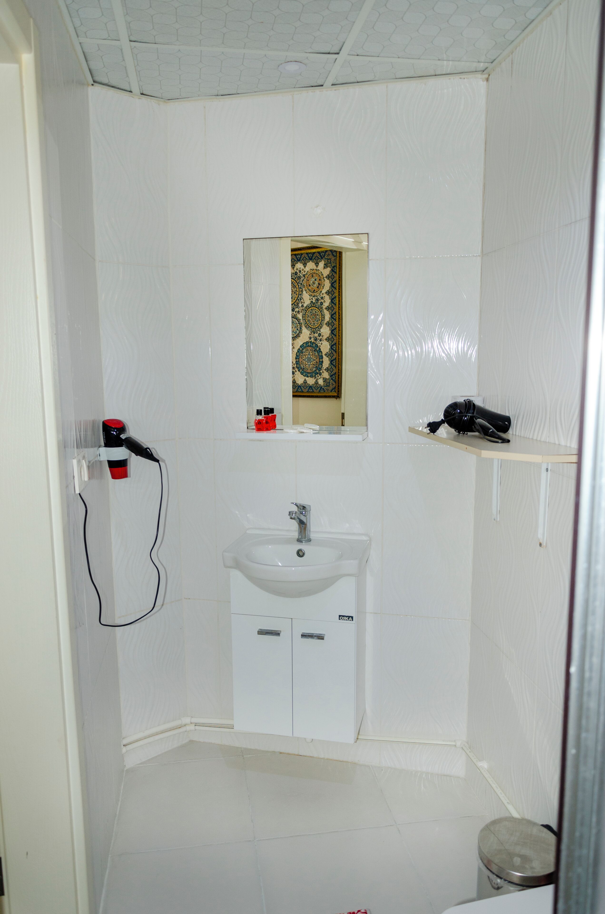 A small, white bathroom with a sink, mirror, and a shelf with toiletries.