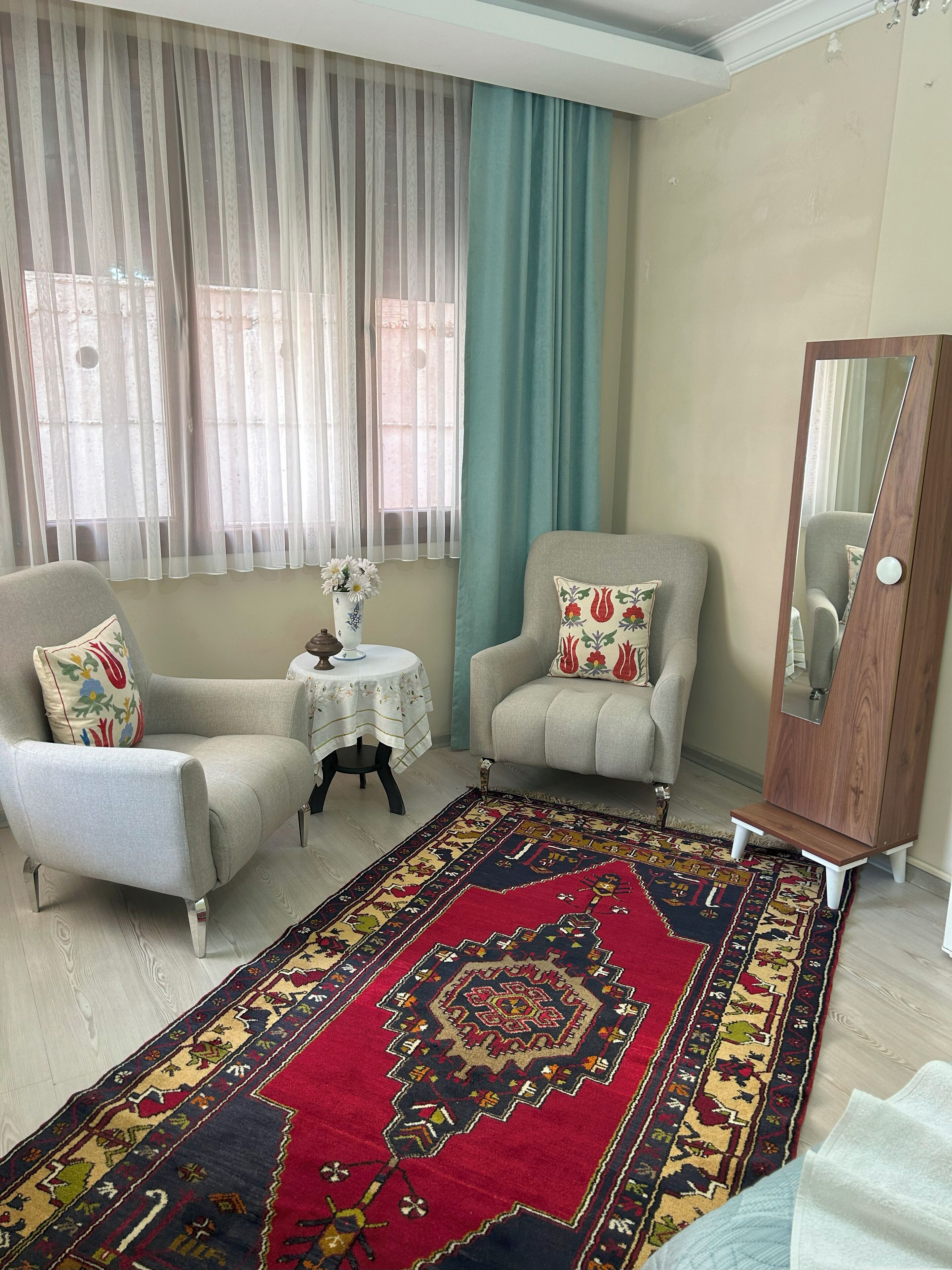 A living room with two armchairs, a small round table with a vase, a large mirror, and a patterned rug.