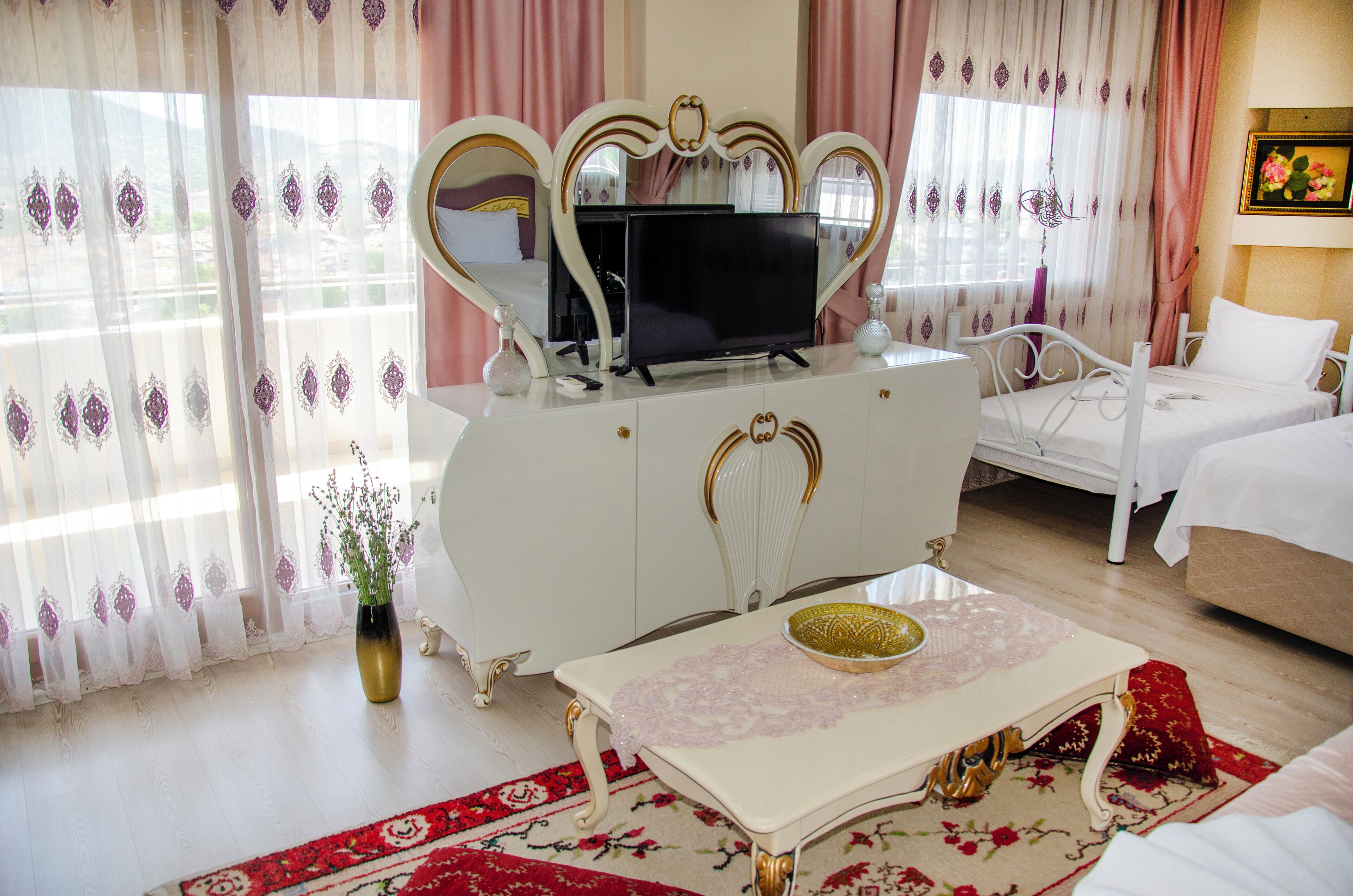 Habitación de hotel con una cama blanca, una mesita de noche con un florero, un aparador con televisor y una alfombra estampada en rojo y blanco.