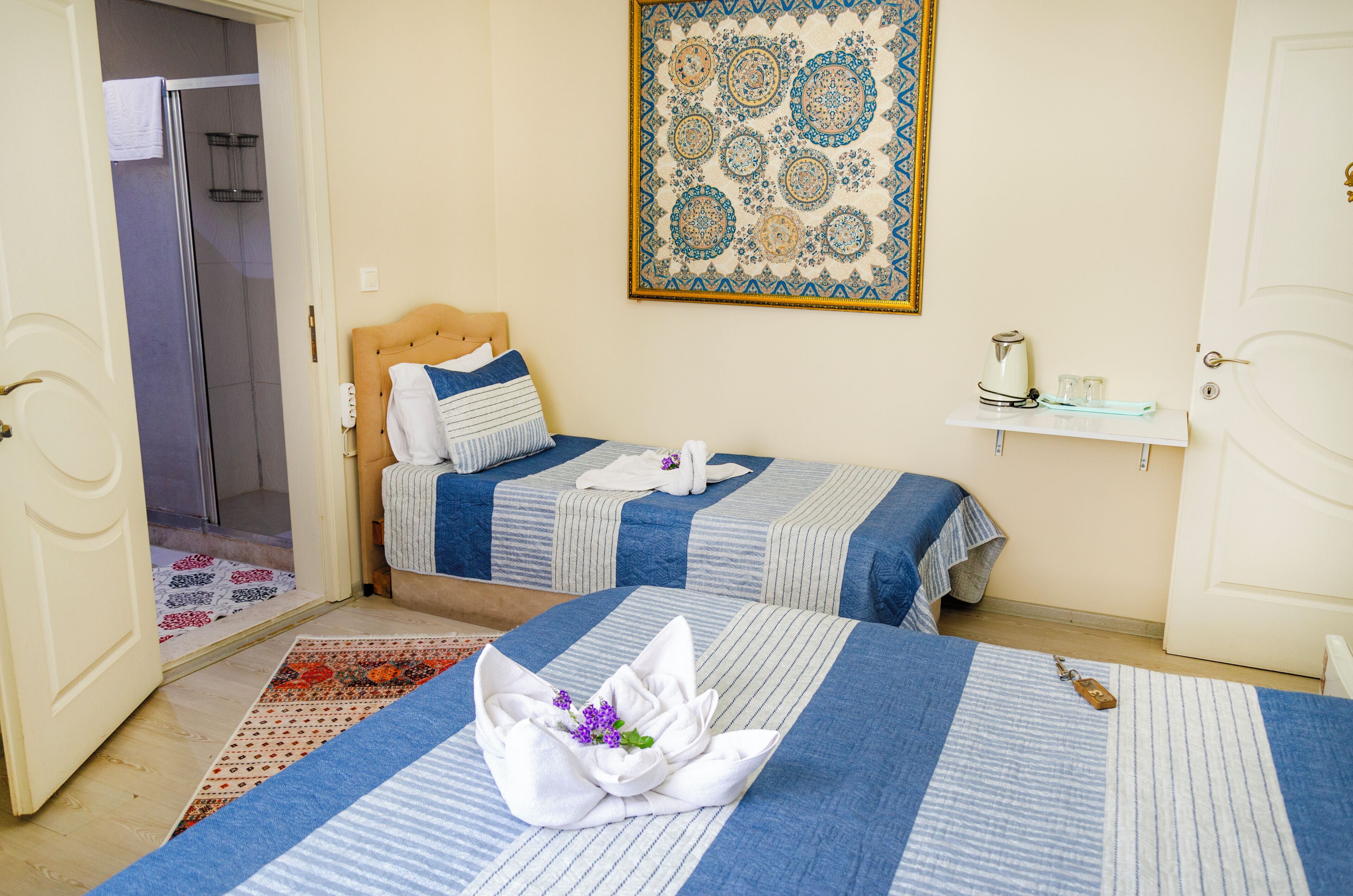 A hotel room with two beds, a shower, a decorative wall hanging, and a small shelf with a kettle and glasses.