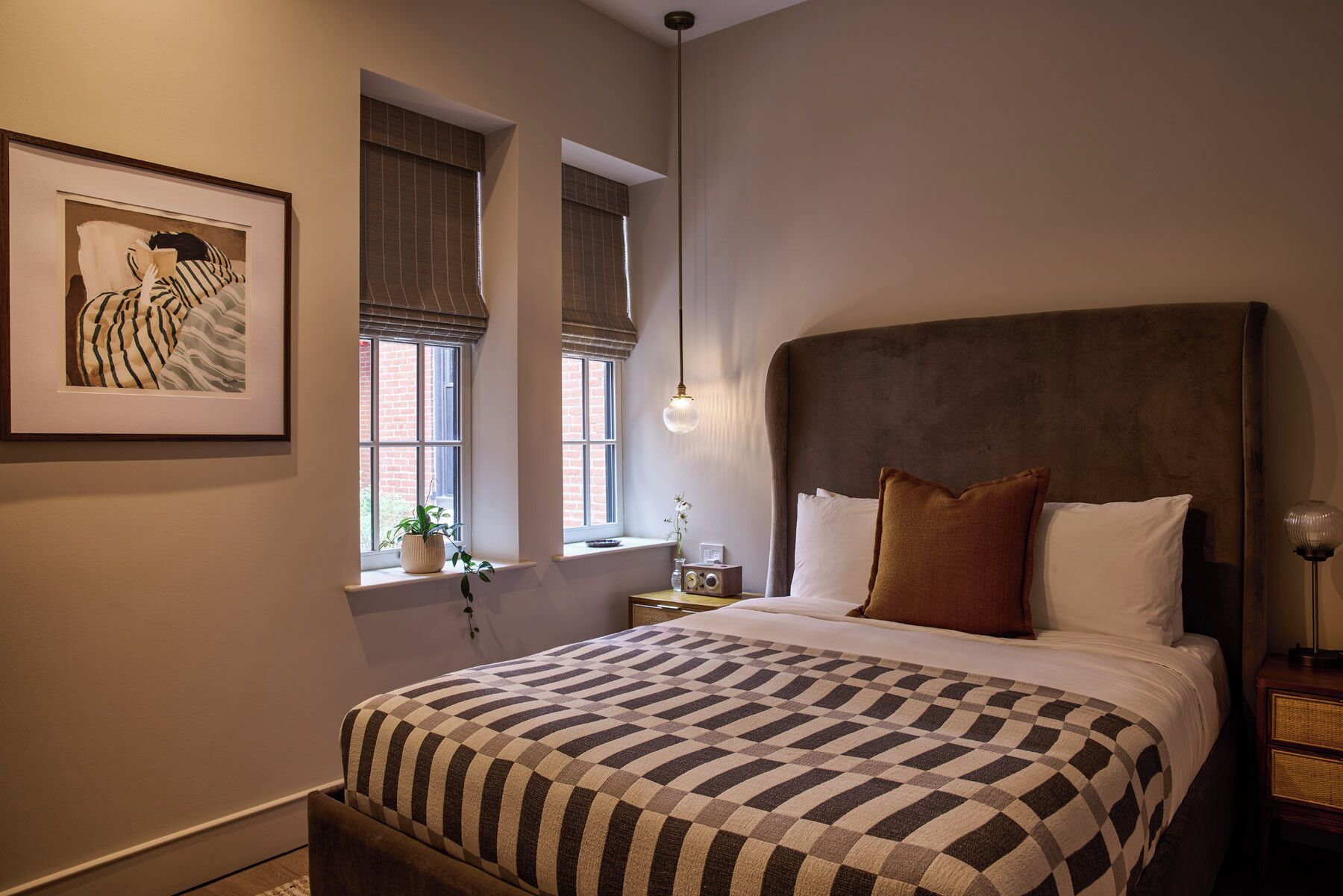 A bedroom with a large bed, a headboard, a window with blinds, a bedside table with a lamp, and a framed picture on the wall.
