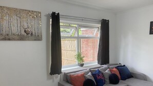 A living room with a grey sectional sofa, a painting of a forest scene, a window with black curtains, and a view of a brick wall and greenery outside. - 4BR Sleeps 9 With Garden and Parking (Birmingham)