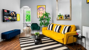 Een moderne woonkamer met een gele bank, een blauwe bankje, een zwart-wit gestreept tapijt, een groene fauteuil en een groot raam.