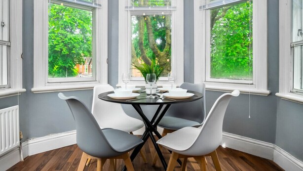 Een eethoek met een ronde tafel, vier stoelen en uitzicht op bomen door grote ramen.