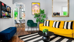 Een moderne woonkamer met een gele bank, een blauwe hocker, een zwarte salontafel en een grote spiegel. Er staat een groene fauteuil, een eethoek met een witte tafel en stoelen, en een groot schilderij aan de muur.