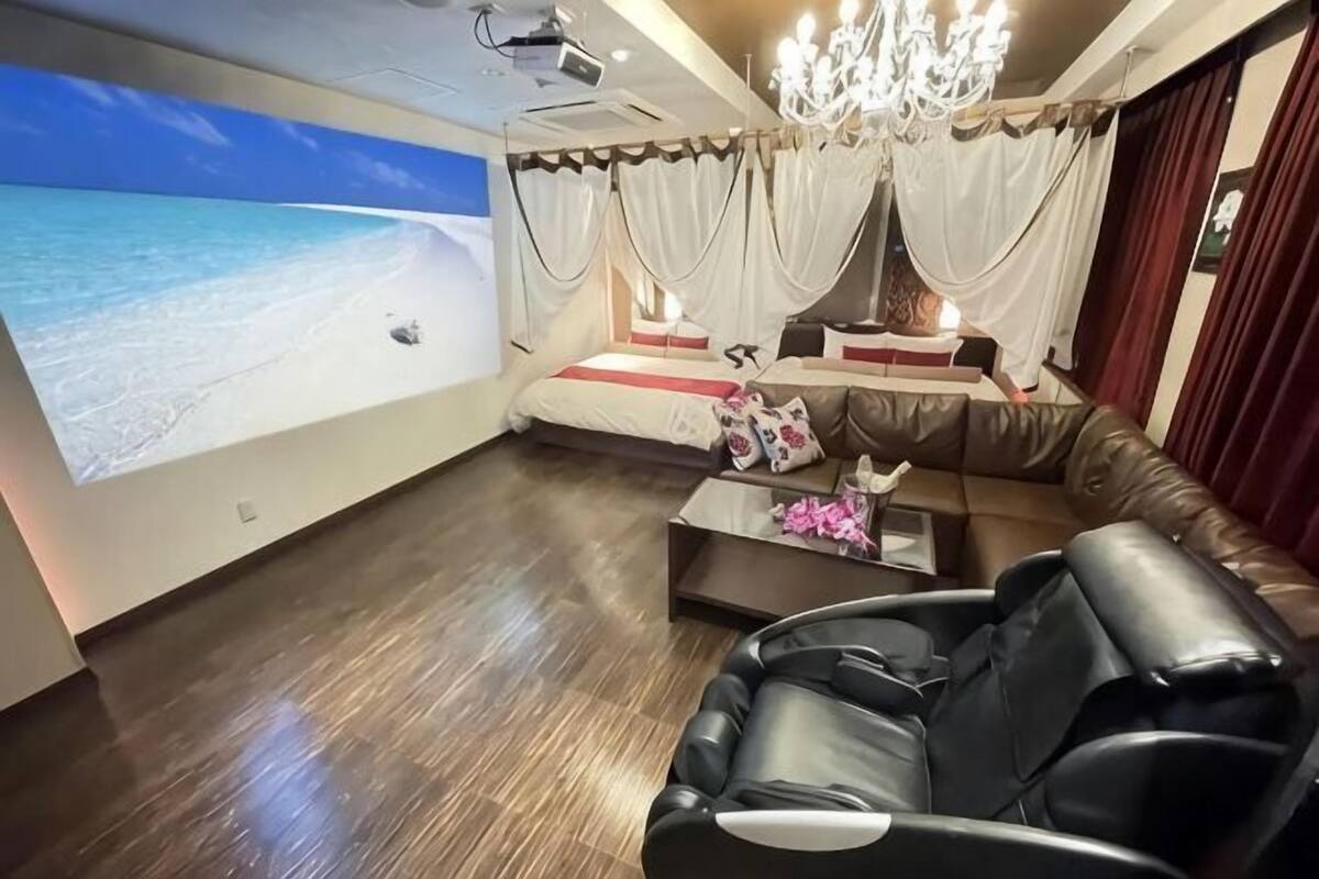 Een kamer met een groot scherm waarop een strand te zien is, een ligbank, een massagestoel en een kroonluchter.