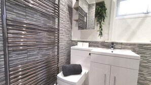 A modern bathroom with a grey tiled wall, a white toilet, a white sink with a mirror above it, and a small window. - Captivating 6-bed House in Havant With Parking (Havant)
