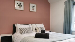 A bedroom with a bed, white bedding, a striped pillow, a dark throw blanket, a nightstand, a drawer, and two framed plant pictures on the wall. - Captivating 6-bed House in Havant With Parking (Havant)