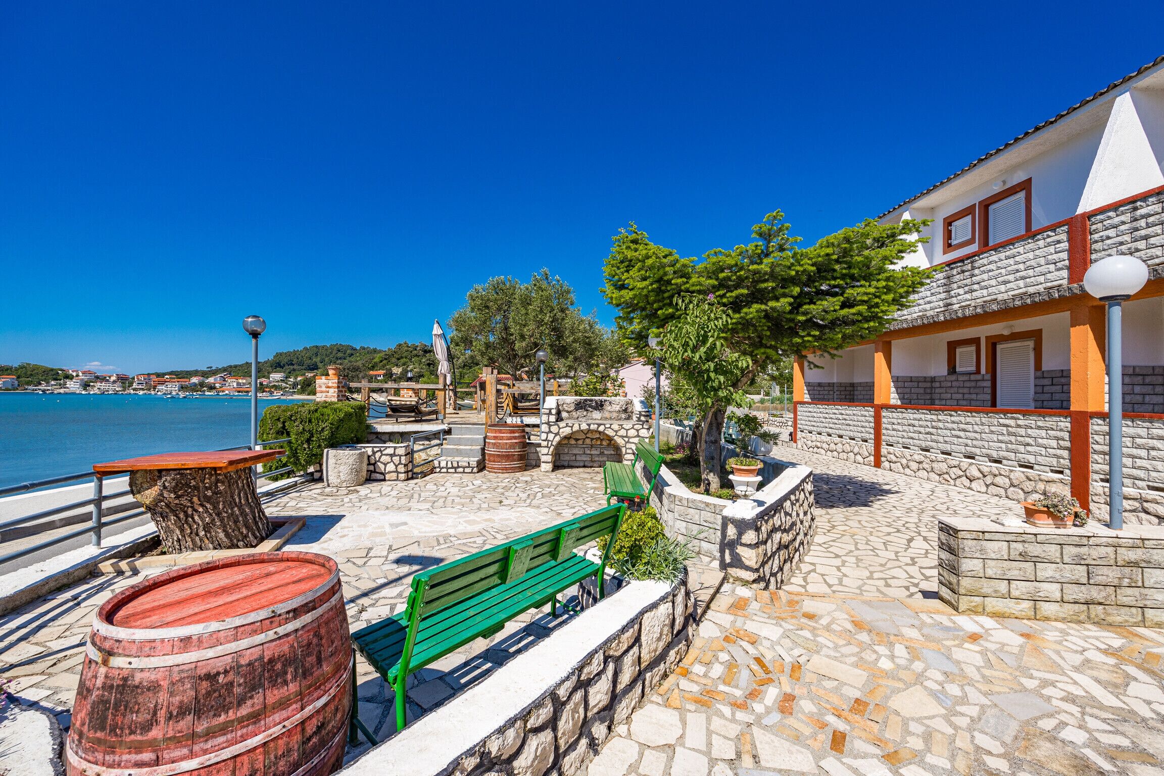 A coastal promenade with stone flooring, a green bench, a wooden barrel, and a building with a tiled roof.
