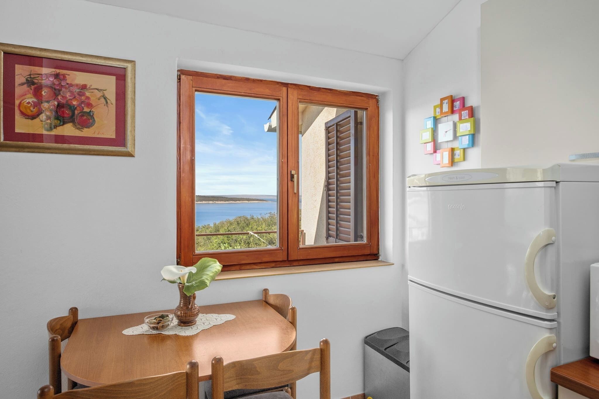 A small kitchen with a wooden table and chairs, a window with a view, a refrigerator, and a framed picture on the wall.