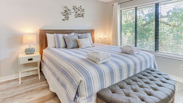 Een slaapkamer met een groot bed, nachtkastje, een bankje en een raam met jaloezieën.