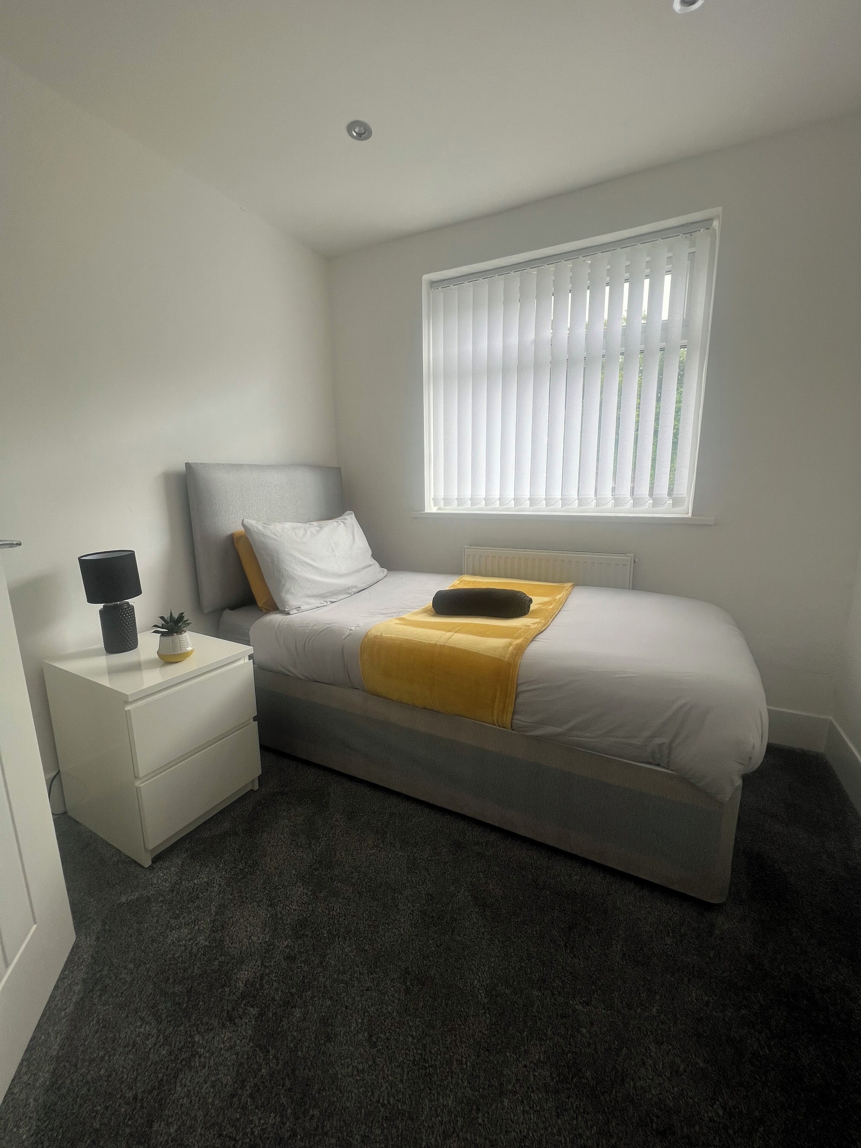 A bedroom with a bed, a bedside table with a lamp, a window with blinds, and a small plant.