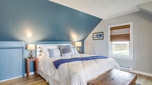 A bedroom with a large bed, a wooden nightstand, a bedside table, a window with blinds, and a picture on the wall. - Unique Tangier Cottage w/ Chesapeake Bay Views! (Tangier)