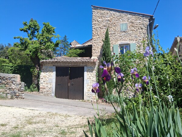 Un bâtiment en pierre avec une porte en bois, entouré de verdure et surplombé d’un ciel bleu clair.