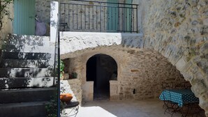 Un patio de piedra con una pequeña mesa y sillas, un balcón con baranda de metal y una escalera que sube a un nivel superior.