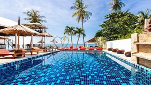 Una piscina con baldosas de mosaico azul, sillas de descanso, cojines rojos, palmeras y vistas al mar.