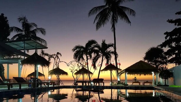 Área de piscina en un resort, con palmeras, sombrillas de paja y vistas al atardecer.