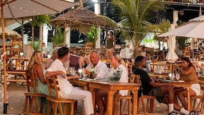 Un espacio de comedor al aire libre con mesas y sillas de madera, rodeado de sombrillas y palmeras.