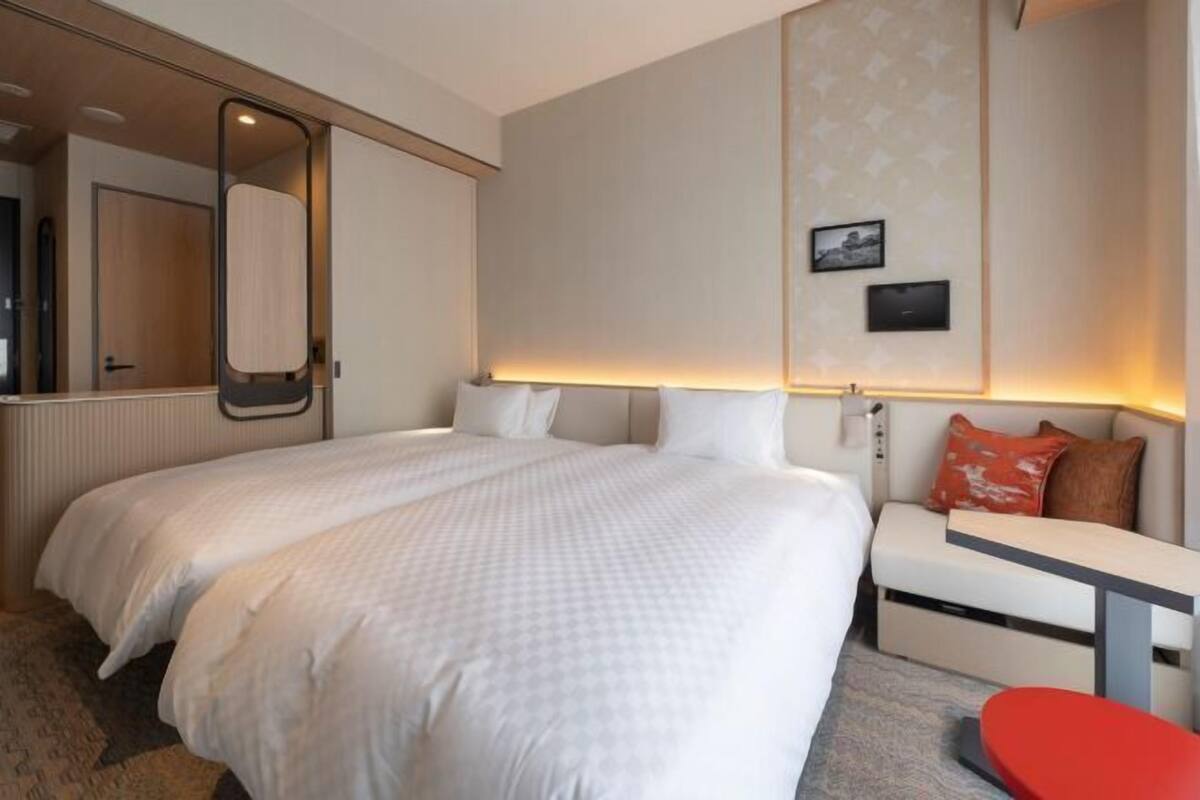 A hotel room with a large bed, a bedside table, a red chair, a mirror, and a wall-mounted picture.