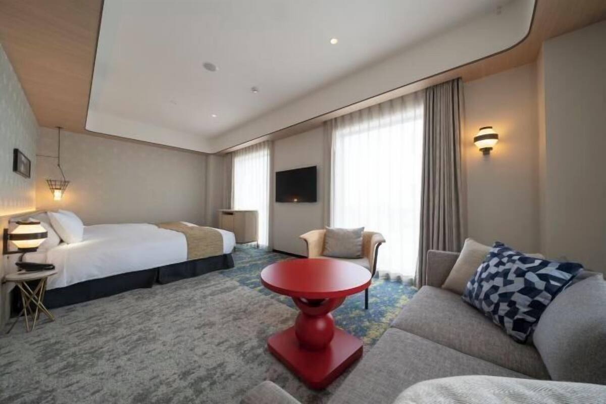 A hotel room with a bed, a sofa, a red table, a TV, and a window with curtains.