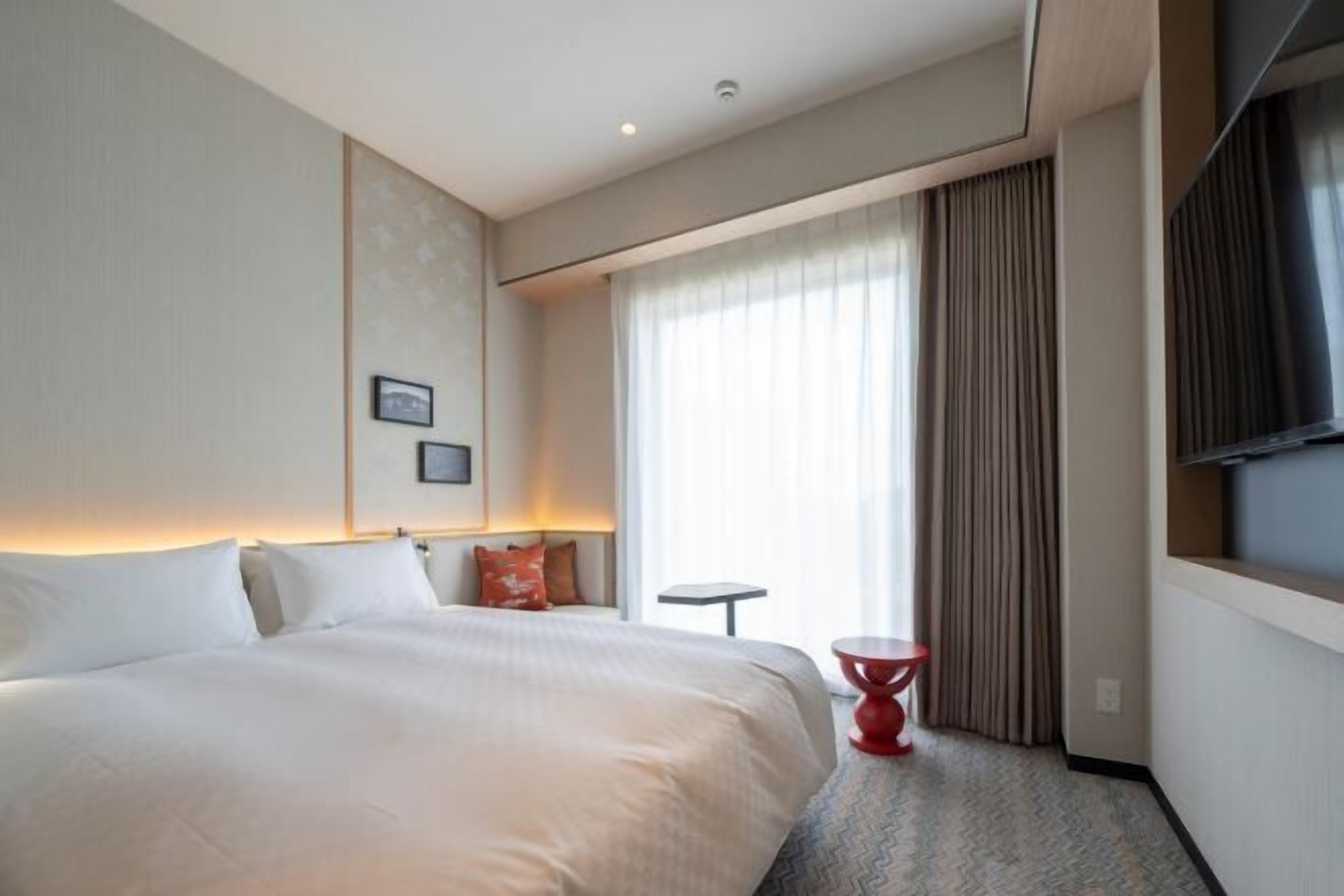 A hotel room with a bed, a small table, a red chair, a TV, and a window with curtains.