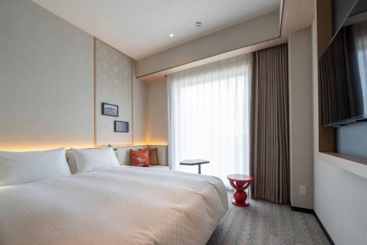 A hotel room with a bed, a small table, a red chair, a TV, and a window with curtains.