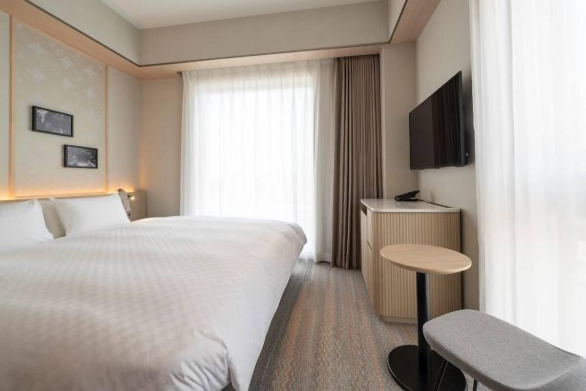 A hotel room with a large bed, a desk, a chair, a TV, and a window with curtains.