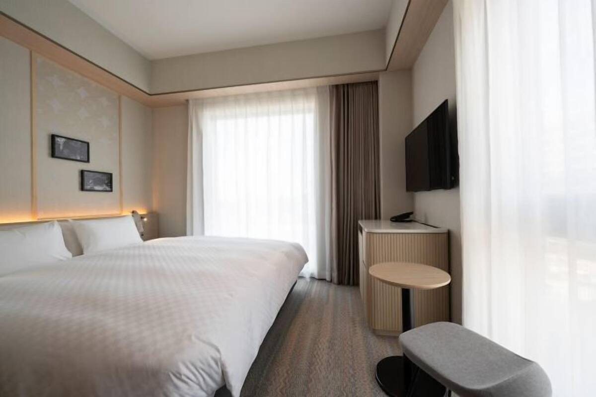 A hotel room with a large bed, a small desk, a chair, a flat-screen TV, and a window with curtains.