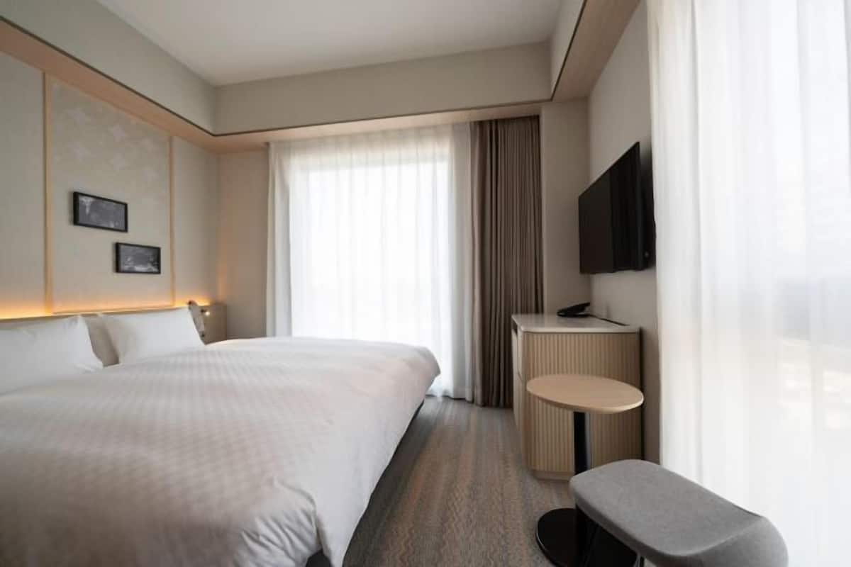 A hotel room with a large bed, a small desk, a chair, a flat-screen TV, and a window with curtains.