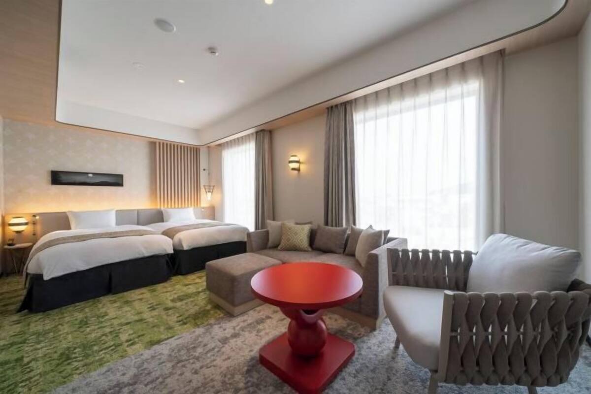 A hotel room with a bed, two bedside tables, a red table, a sofa, and a chair.