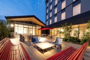 An outdoor seating area with tables and chairs, a wooden deck, and a building with large windows. - Hotel μ Style Inuyama Experience (Inuyama)