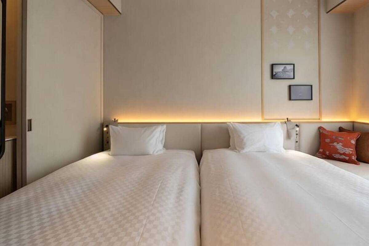 A hotel room with two beds, a headboard with a light, and a wall-mounted picture.