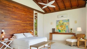 A bedroom with a wooden wall, a bed, a bench, a wicker chair, a lamp, and a painting. - Fazenda São Francisco do Corumbau (Prado)