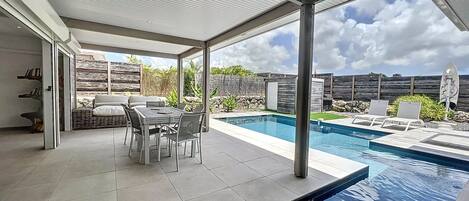 A covered outdoor area with a pool, a dining table with chairs, and a view of the backyard.