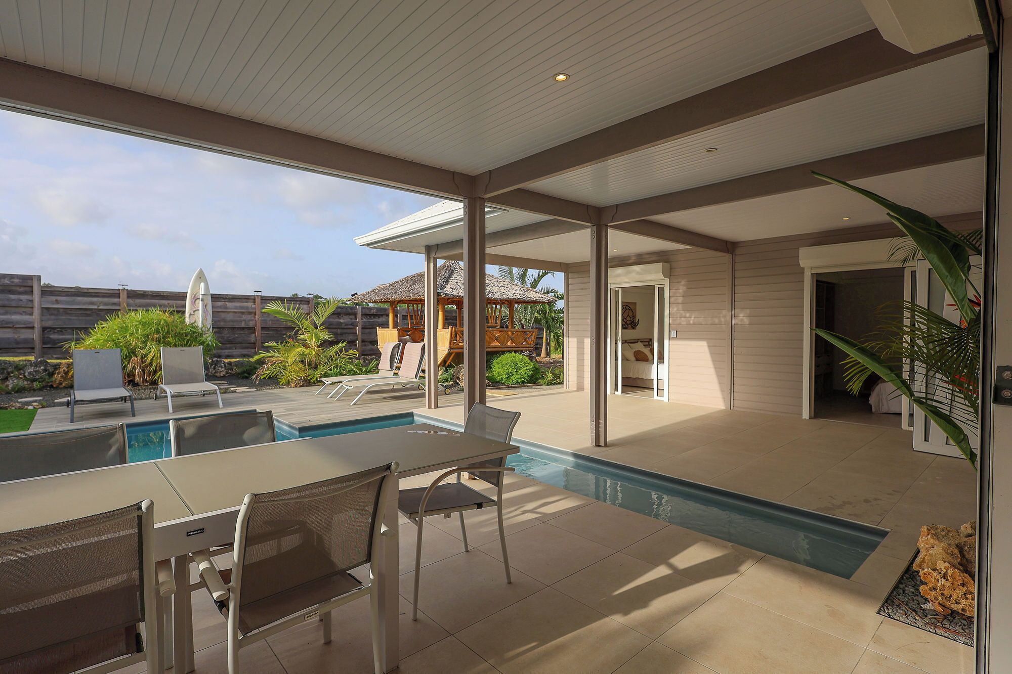A modern outdoor pool area with a table and chairs, a covered seating area, and a view of the backyard.