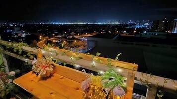A balcony with a wooden table and chairs, decorated with flowers and a lantern. A cityscape is visible in the background.